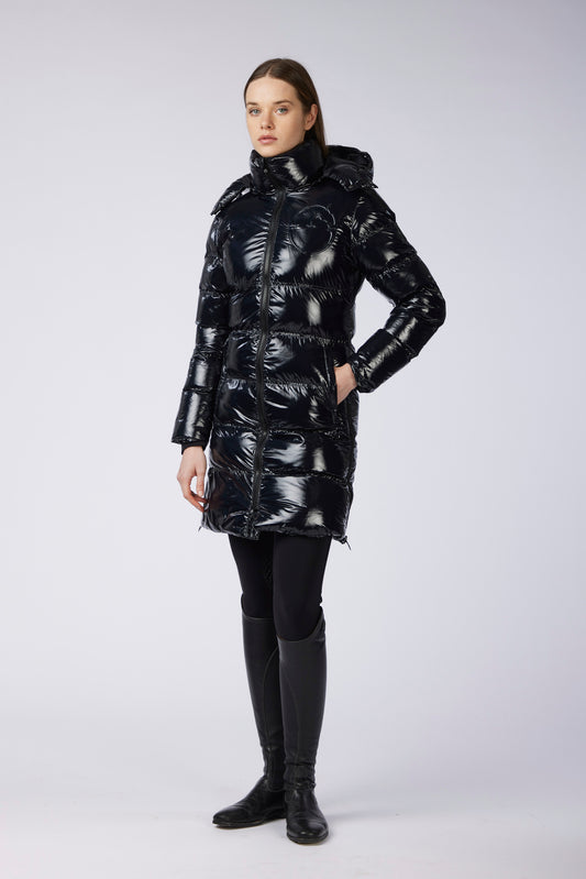 A woman modeling a shiny black puffer coat with a high collar and hood, paired with black leggings and black boots, against a light gray studio background.
