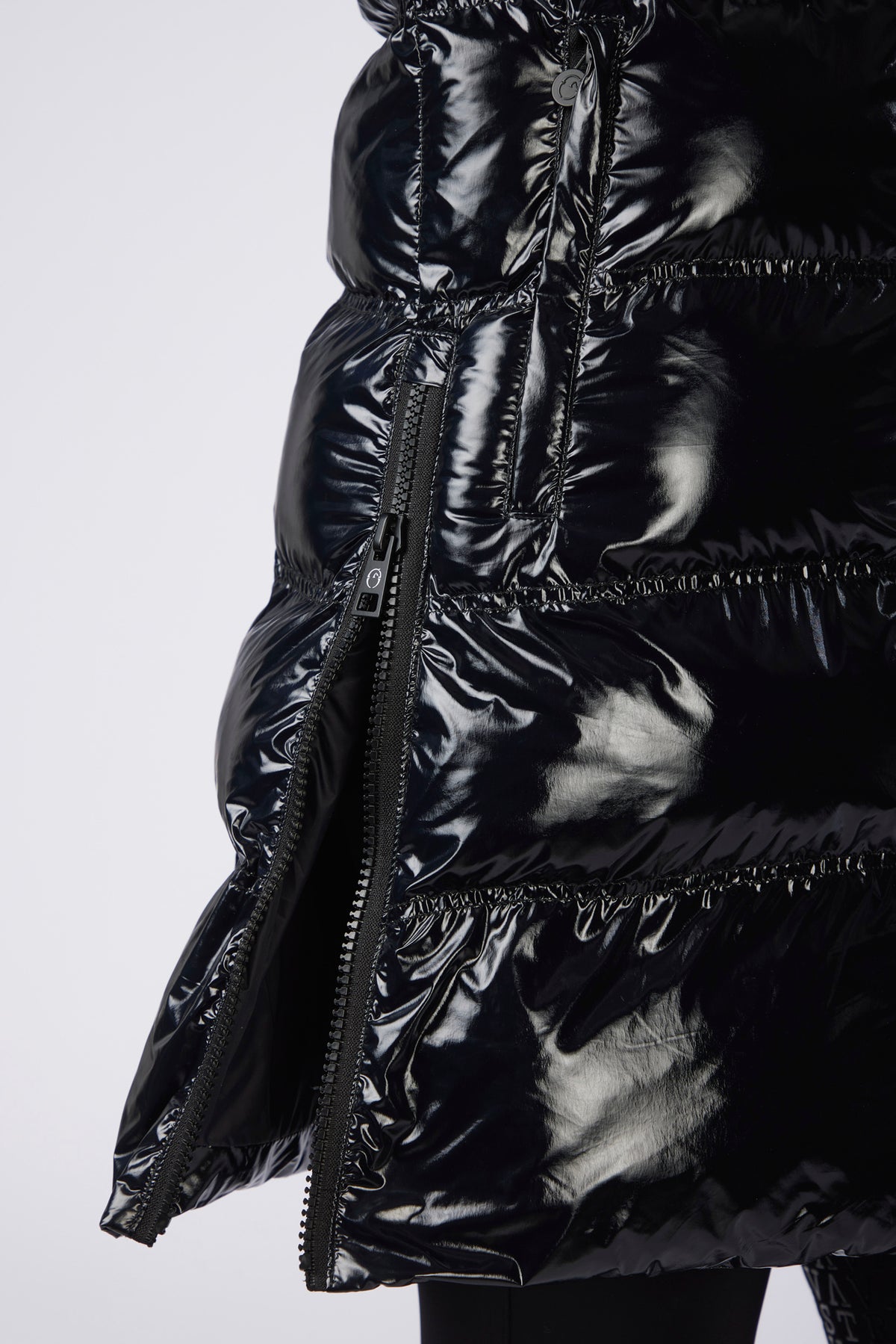 Close-up of a glossy black puffer jacket with horizontal quilting, a front zipper, and a ruffled hem detail, shown against a light gray background.