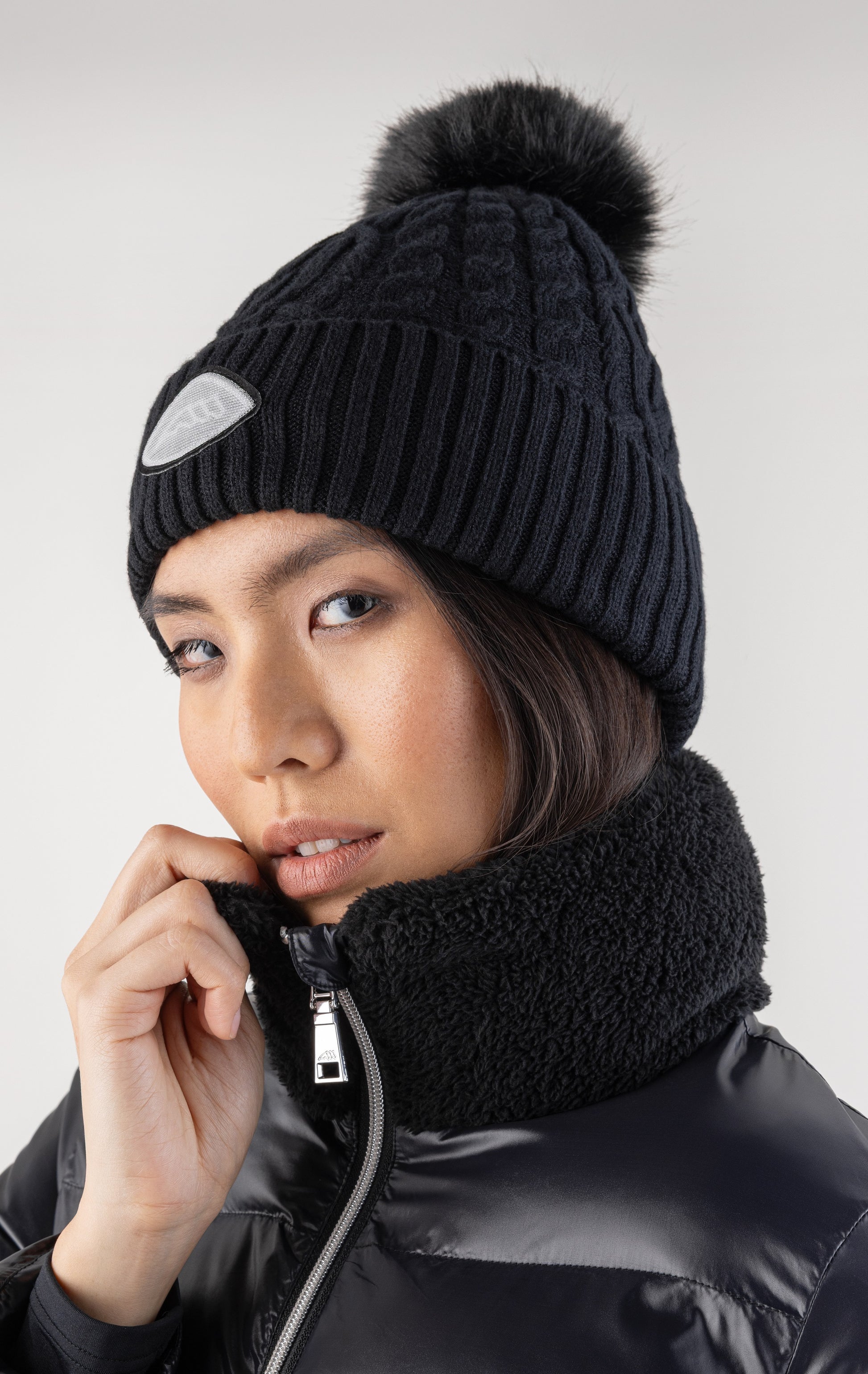 Person wearing a black knit beanie with a pom-pom and black jacket on a white background