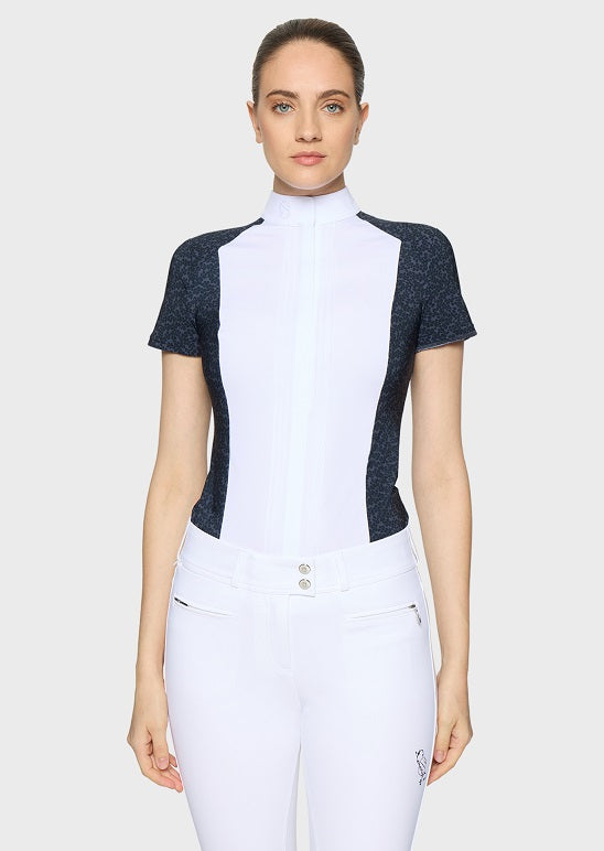 A woman modeling a white and dark gray color-blocked short-sleeve equestrian shirt with a high mock neck and white riding pants against a light gray background.