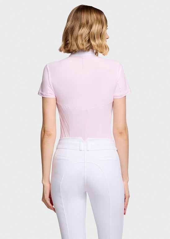 A woman shown from the back wearing a light pink short-sleeve mock-neck equestrian shirt and white riding breeches against a light gray studio background.