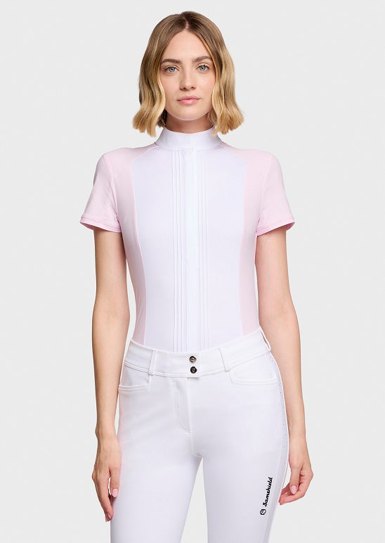 A woman modeling a white and light pink short-sleeve equestrian riding shirt with a high mock neck, paired with white riding pants, against a light gray studio background.