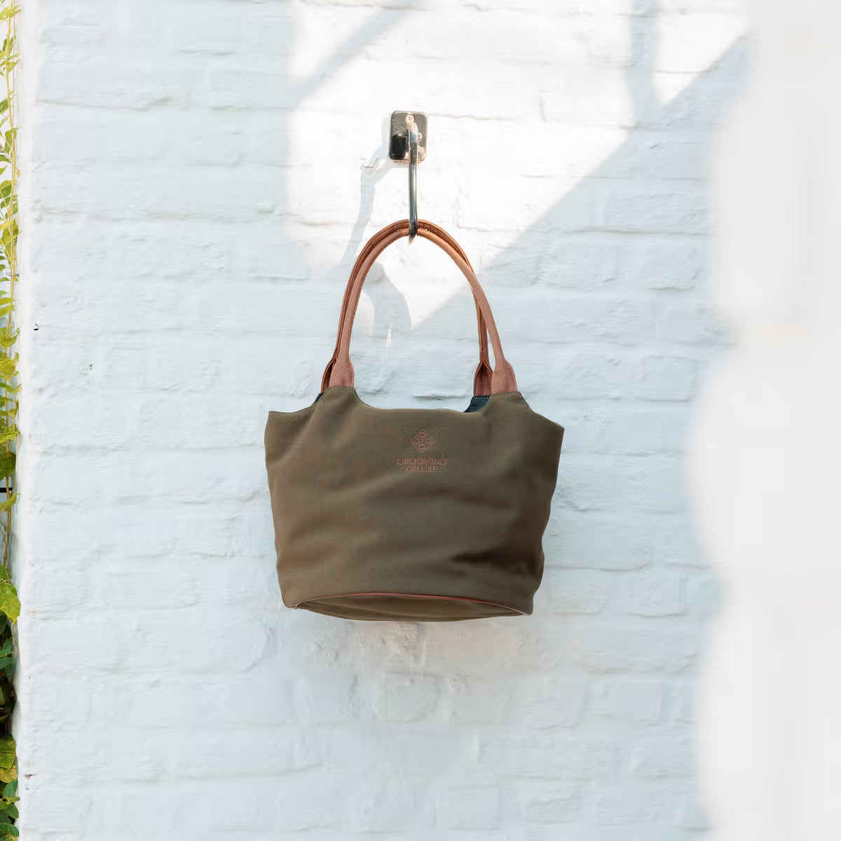 Green handbag with brown handles hanging on a white brick wall.