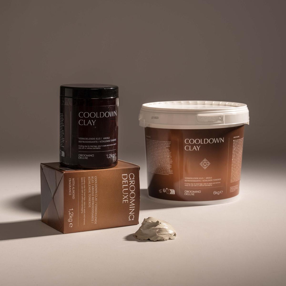 Two containers of Cooling Down Clay grooming product on a neutral studio background, with a small pile of the clay product in front. The set includes a black jar on a brown box and a larger white-lidded tub with a brown label.