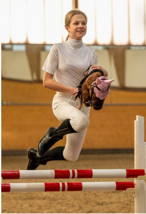 Girl in equestrian attire preparing to jump over a red and white obstacle on her hobby horse in an indoor arena.