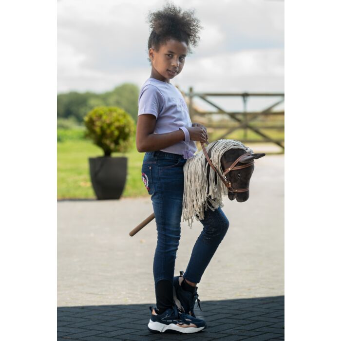 Young girl holding a toy horse outdoors on a paved area with greenery in the background.