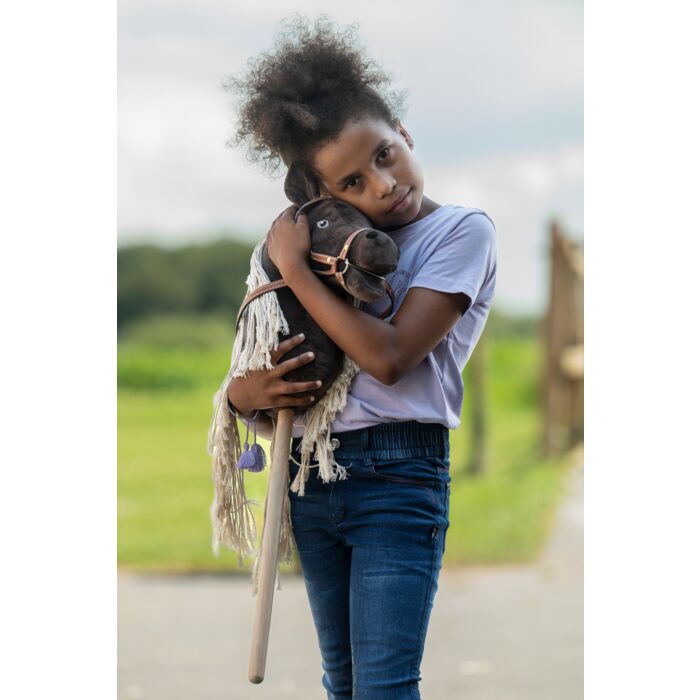 A young girl with curly hair stands outdoors on a path, holding a dark brown wooden hobby horse with a fringed mane and leather reins.