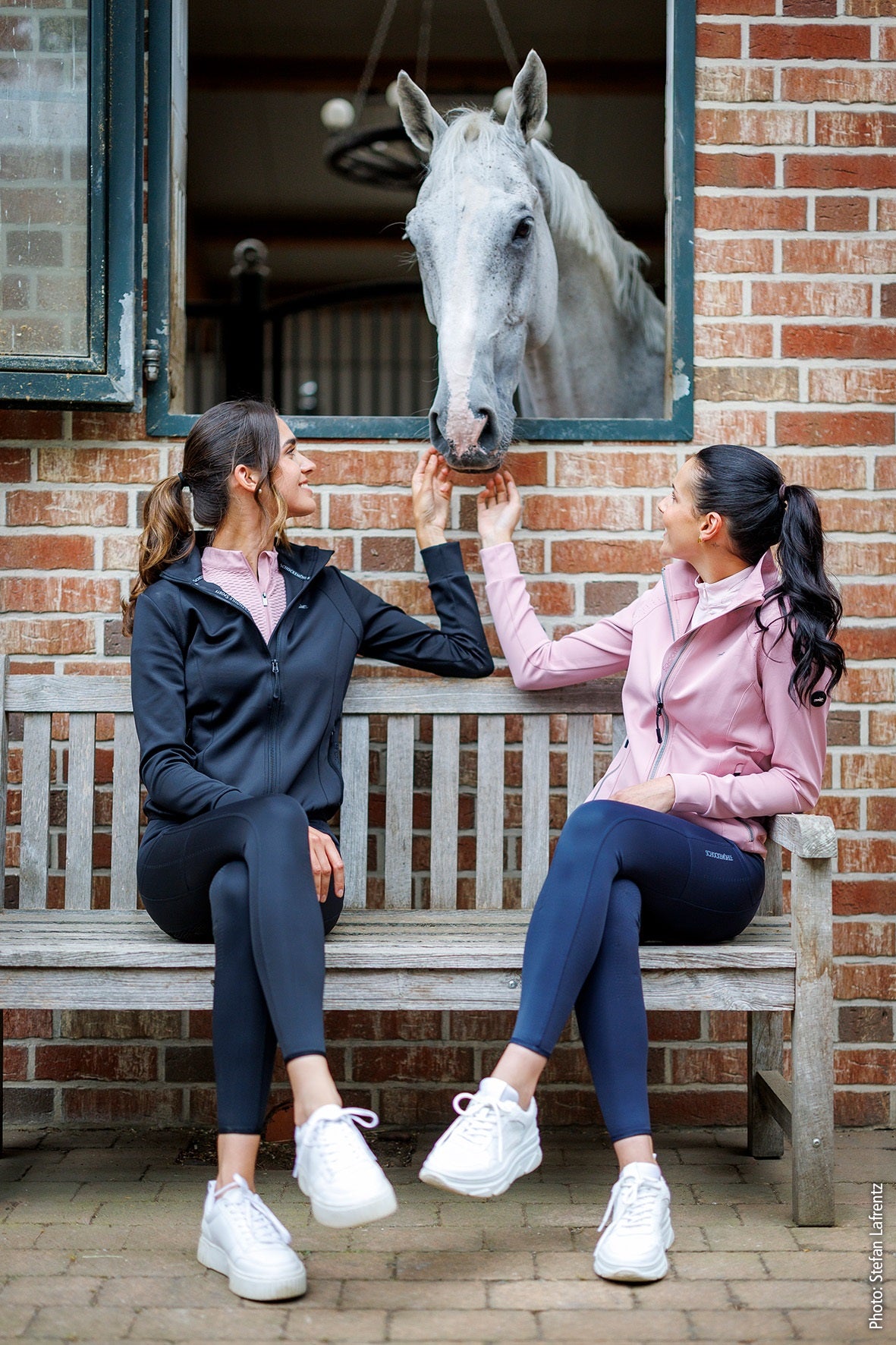 Two women sitting on a bench with a horse looking out of a stable door.