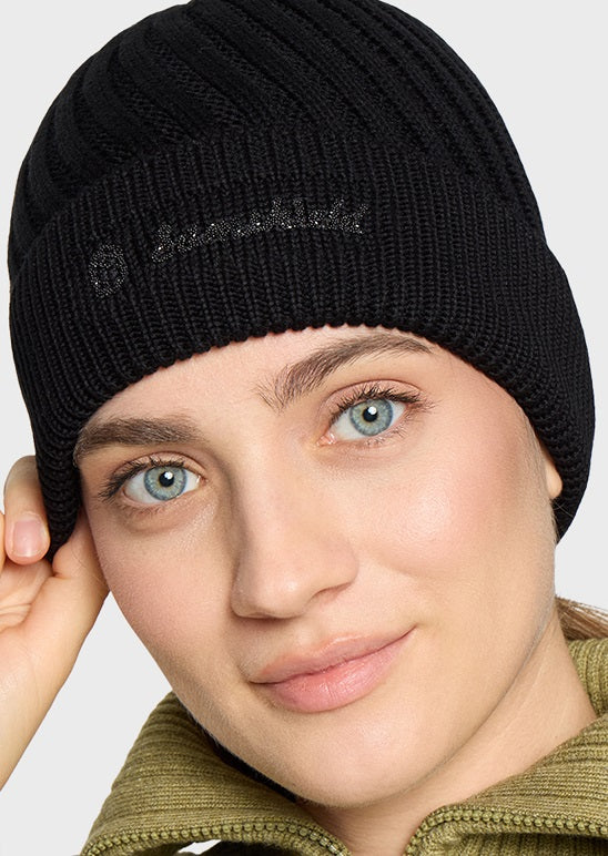 A close-up of a woman wearing a black ribbed knit beanie with a subtle embroidered logo on the front, paired with an olive green zip-up jacket against a light gray background.