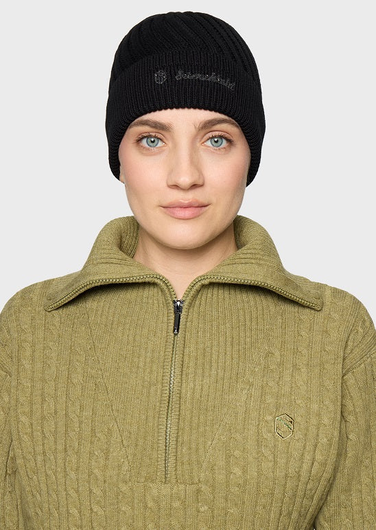 A woman wearing a black ribbed knit beanie with a small white embroidered logo and an olive green ribbed quarter-zip sweater, photographed against a light gray studio background.