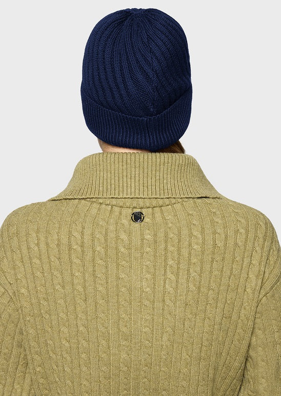Back view of a person wearing a navy blue ribbed knit beanie and a light olive green cable-knit turtleneck sweater against a light gray background.