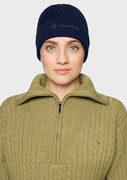 A woman wearing a navy blue ribbed knit beanie with gold embroidered branding and an olive green ribbed knit quarter-zip pullover sweater, shown against a light gray studio background.