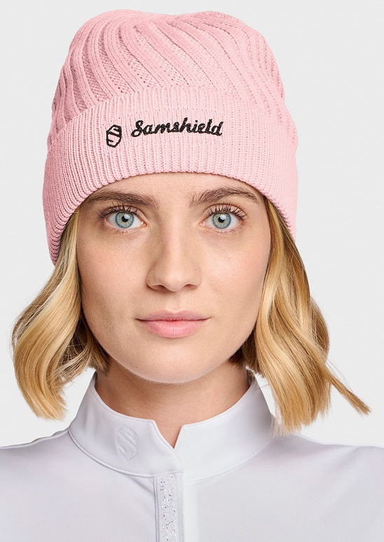 A close-up of a woman wearing a light pink ribbed knit beanie with the black embroidered Samshield logo on the front, paired with a white high-collar top against a light gray background.