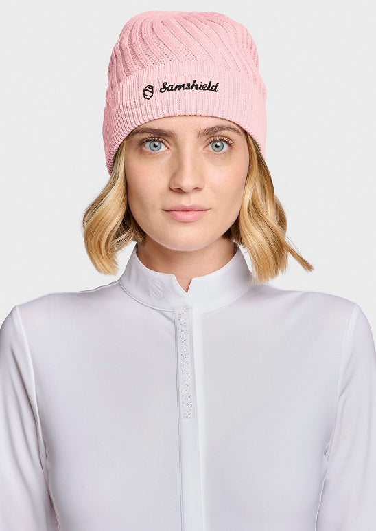 A woman wearing a light pink ribbed knit beanie with 'Samshield' embroidered in black on the front, paired with a white high-neck long-sleeve top against a light gray studio background.