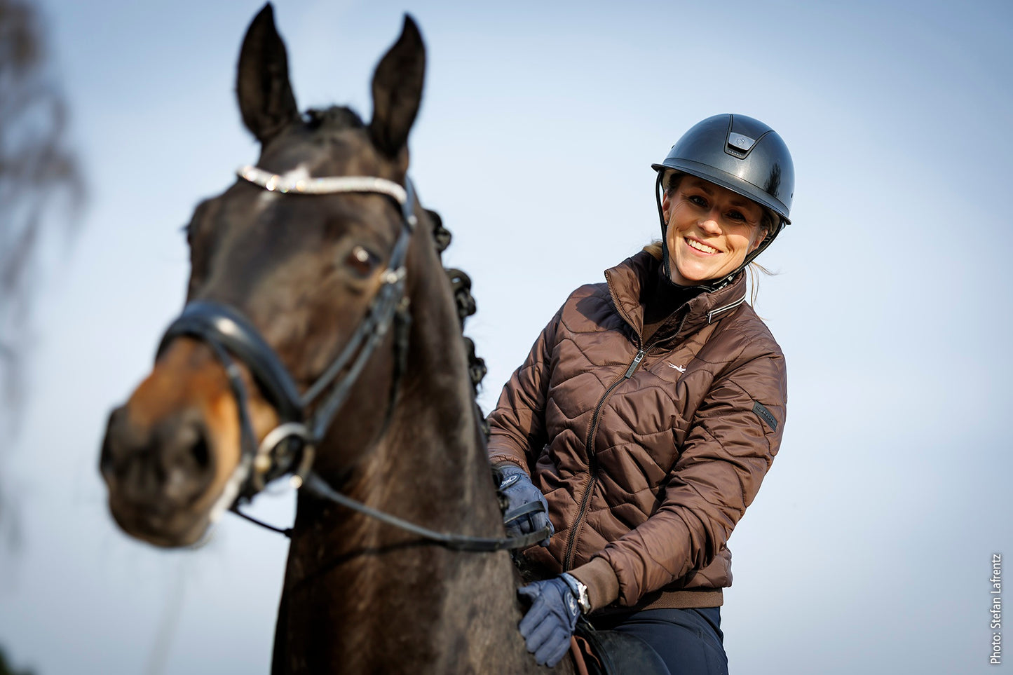 A woman wearing a black riding helmet and a brown quilted jacket sits on a dark brown horse against a pale blue sky.