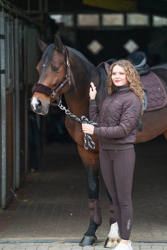 A woman in a brown quilted jacket and matching leggings stands beside a brown horse in a stable, holding the horse's lead rope. The horse has a dark brown saddle blanket and a black saddle.