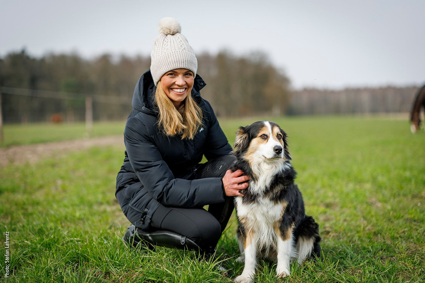 Woman wearing winter clothes kneeling next to a dog in a grassy field