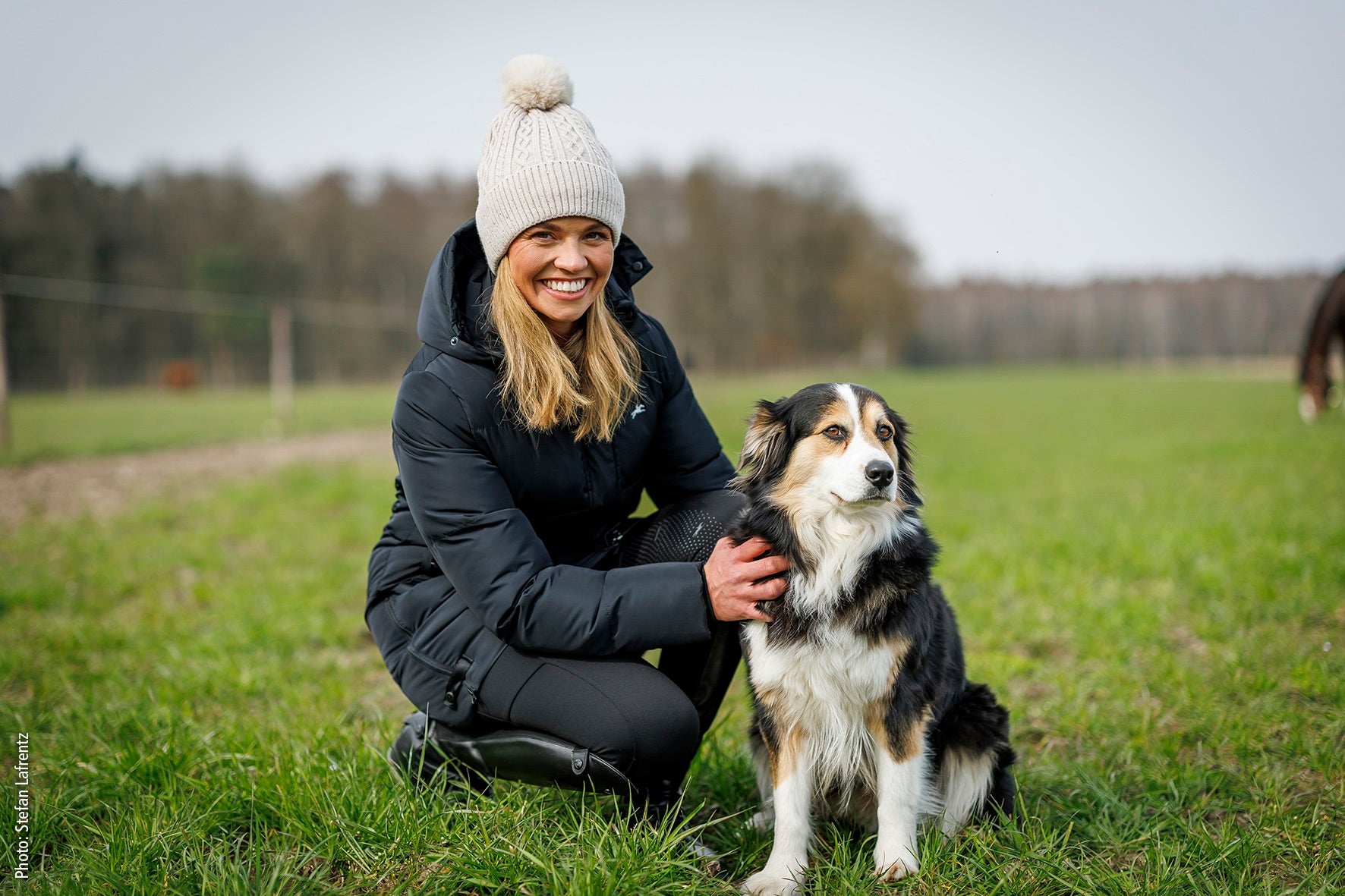 Woman wearing winter clothes kneeling next to a dog in a grassy field