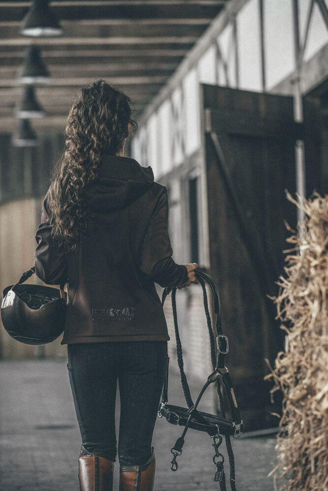 A woman seen from behind in a barn, wearing a dark brown equestrian jacket and holding a bridle and a helmet.