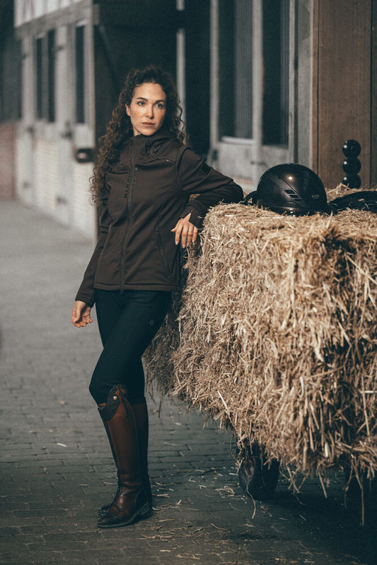 A woman in a dark brown riding jacket, black riding pants, and brown riding boots stands next to a large bale of hay with a black riding helmet on a street in a rustic setting.