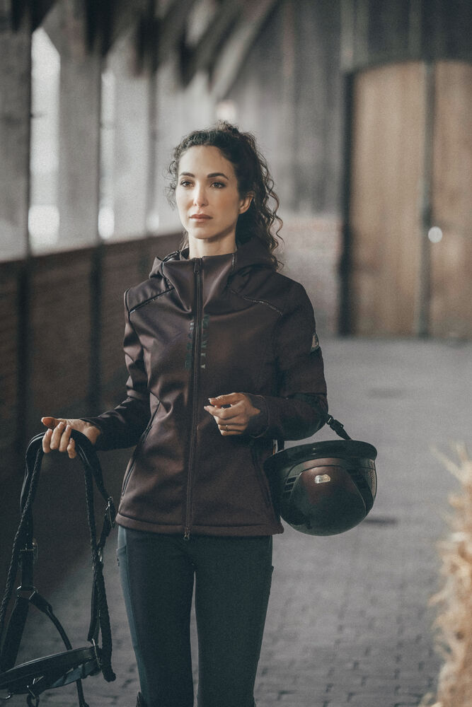 A woman wearing a dark brown equestrian jacket and black pants stands in a rustic stable holding a black riding helmet and equestrian bridle.