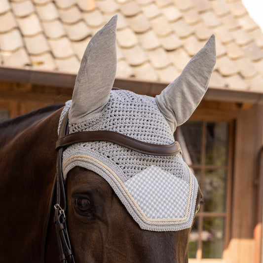 Horse wearing a textured gray fly mask with ear flaps in front of a wooden building.