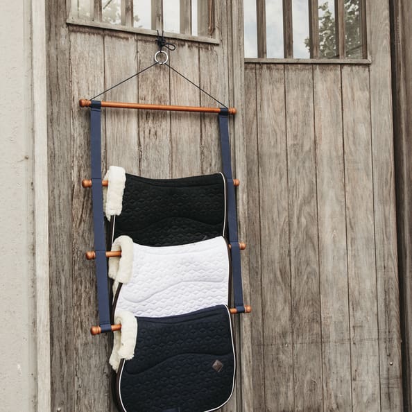 A navy bamboo saddle pad holder rack hanging against a wooden background, displaying four saddle pads.