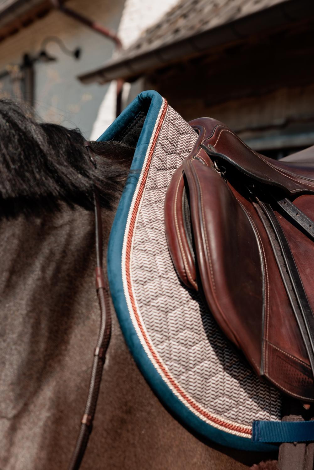 Close-up of a horse wearing a brown saddle with a textured pad on a blurred background