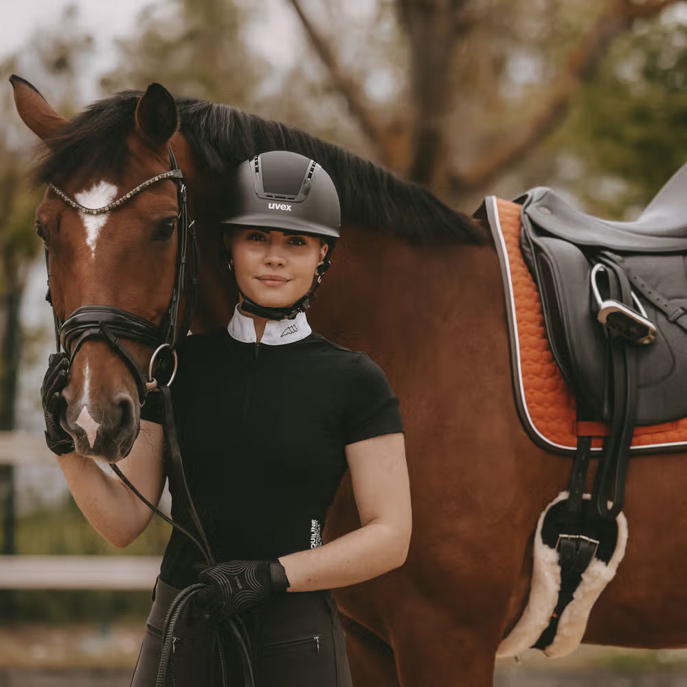 Woman in equestrian gear standing next to a brown horse with a saddle in an outdoor setting.
