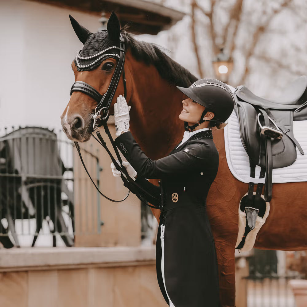 Woman in equestrian attire standing next to a brown horse with a saddle in an outdoor setting.