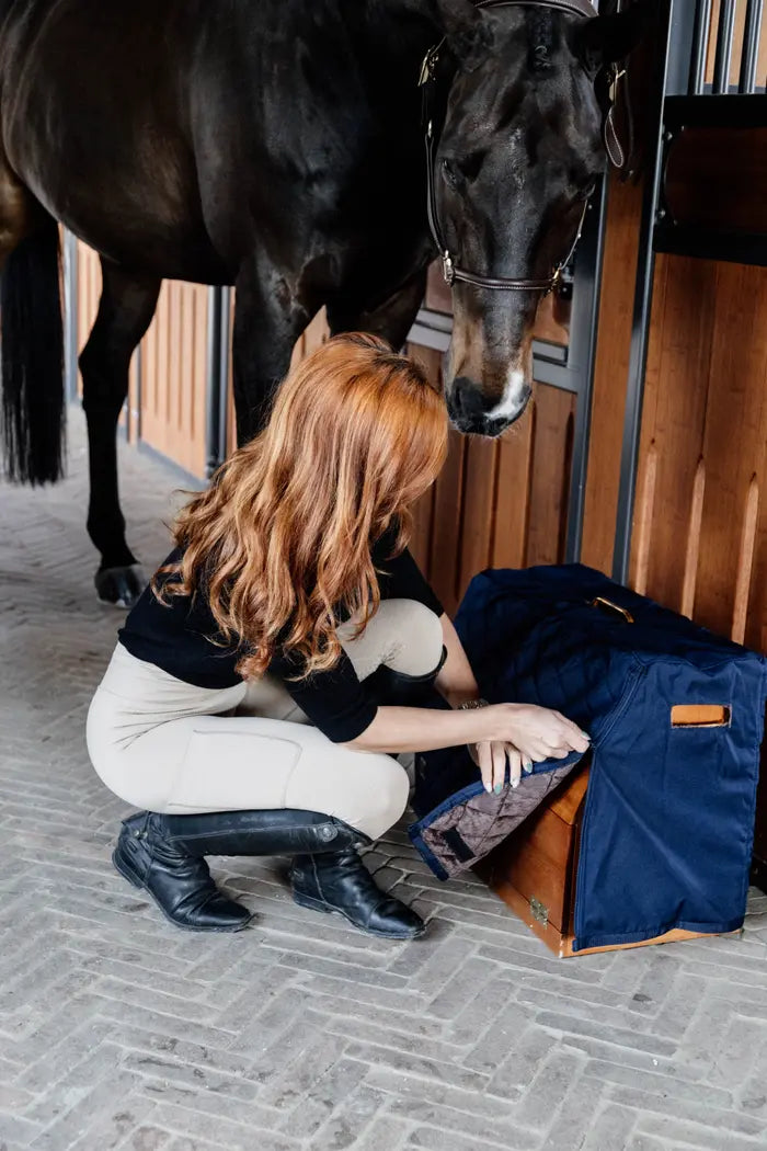 person wearing a jumper and jodhpurs closing a box cover with a horse behind in a stable yard