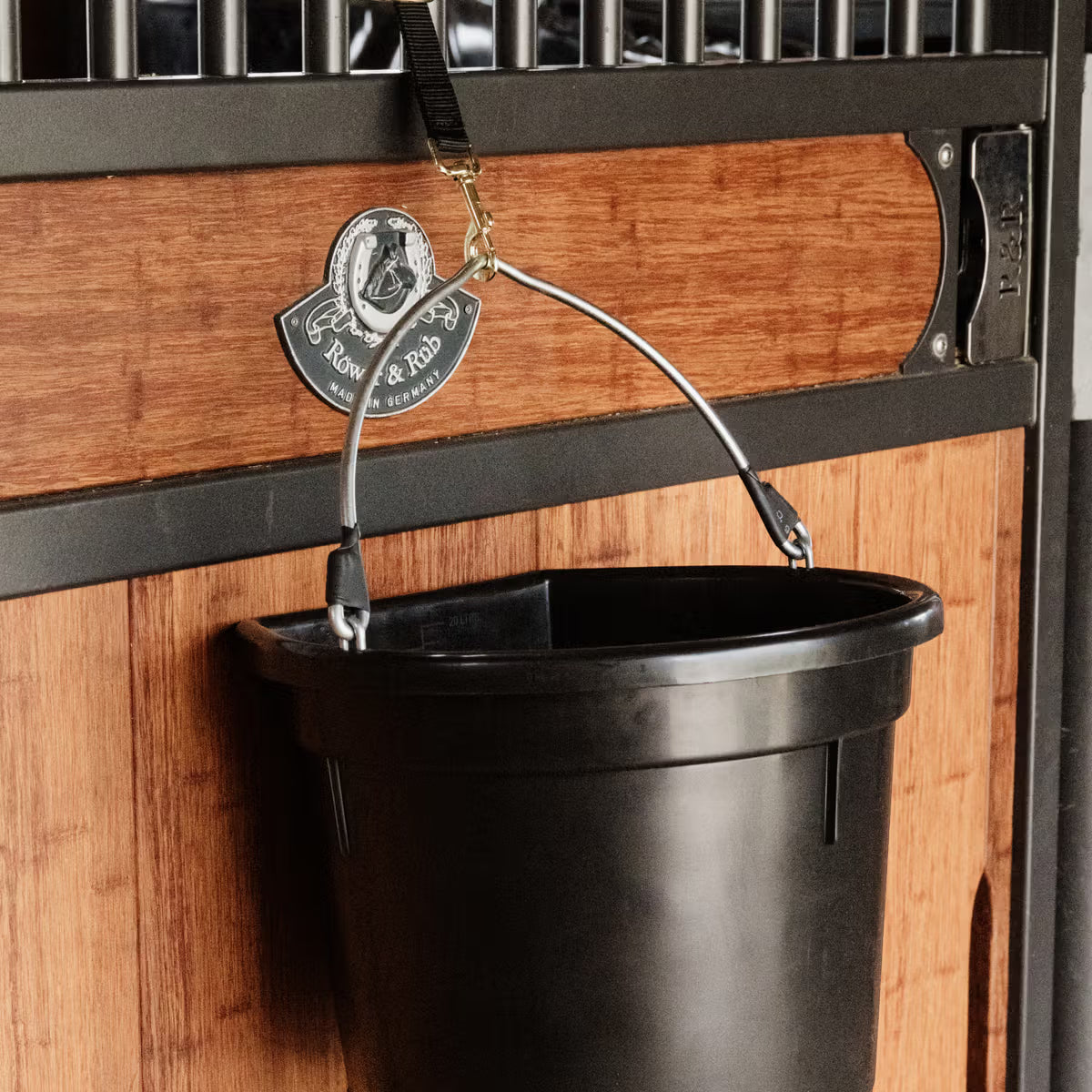 Black bucket hanging from a metal hook on a wooden wall with a brand logo.