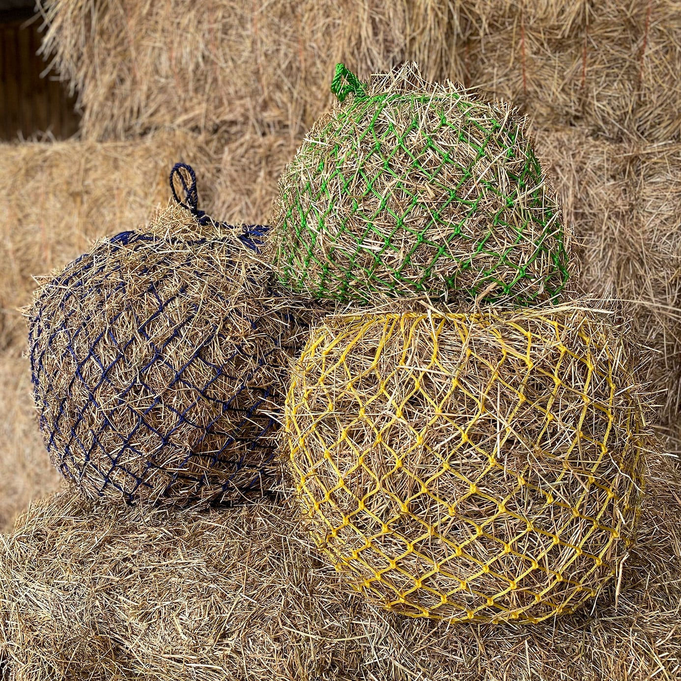 Three round hay nets filled with straw, colored green, yellow, and dark blue, resting on a pile of dry hay.
