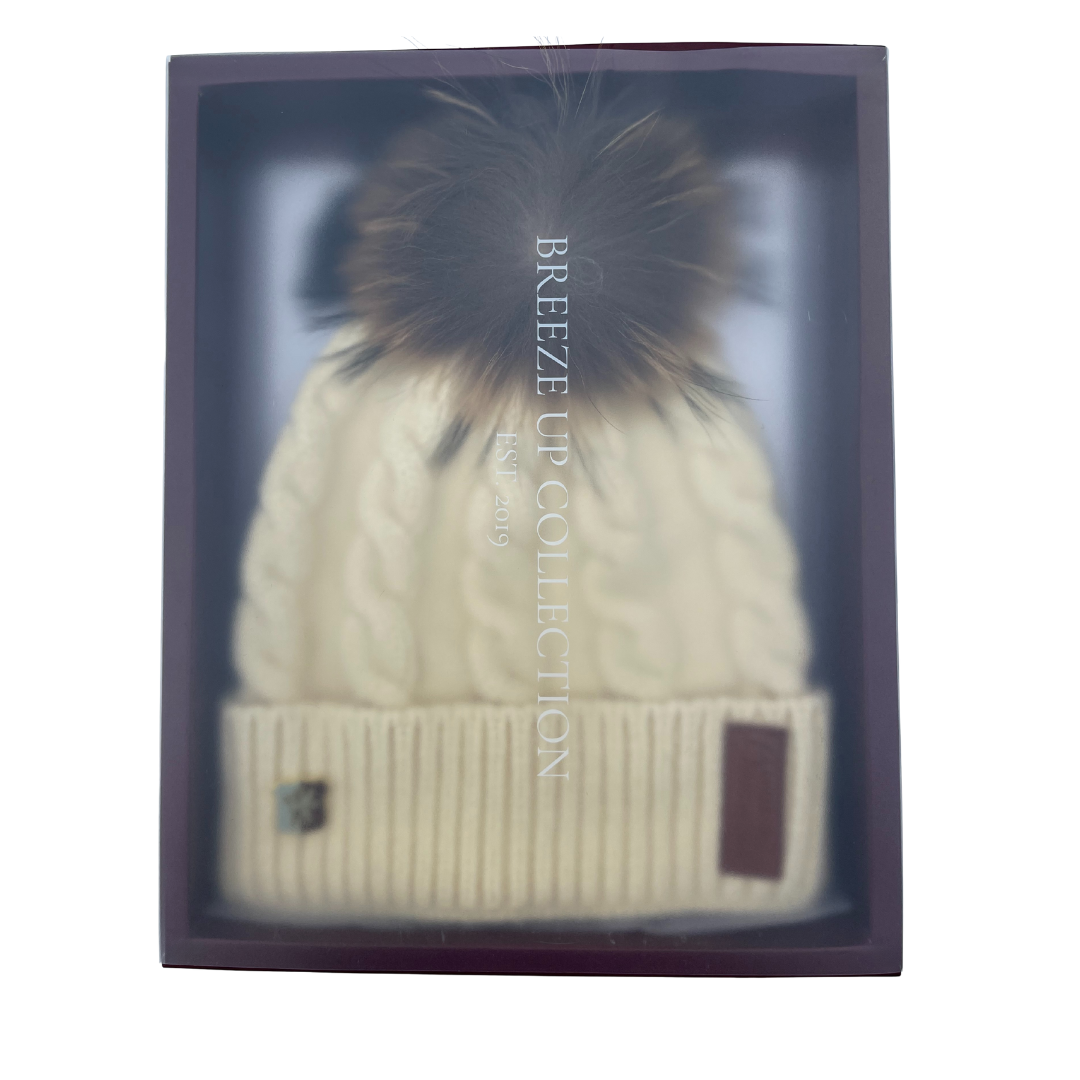 Beige knit beanie with fur pom-pom in a box labeled 'Breeze Up Collection'.