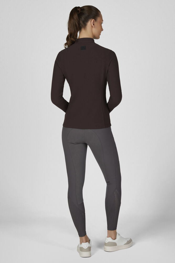 A woman is shown from the back wearing a dark brown long-sleeve athletic top and dark gray leggings, paired with white sneakers, against a light gray studio background.