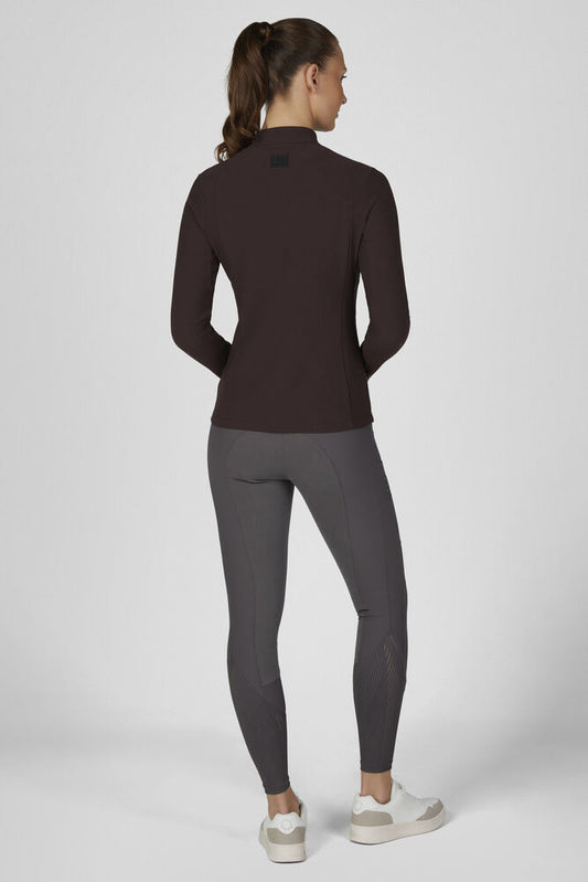 A woman is shown from the back wearing a dark brown long-sleeve athletic top and dark gray leggings, paired with white sneakers, against a light gray studio background.
