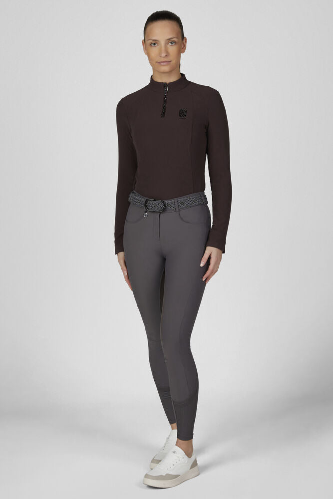 A model stands against a light gray studio background wearing a dark brown long-sleeve quarter-zip top, dark gray fitted riding breeches with a textured gray belt, and white sneakers.