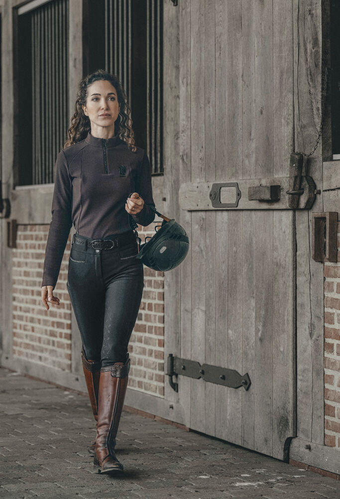 A woman in equestrian attire, including a dark brown mock-neck riding shirt, black riding pants, and brown riding boots, walks alongside a rustic wooden stable door while holding a dark green riding helmet.