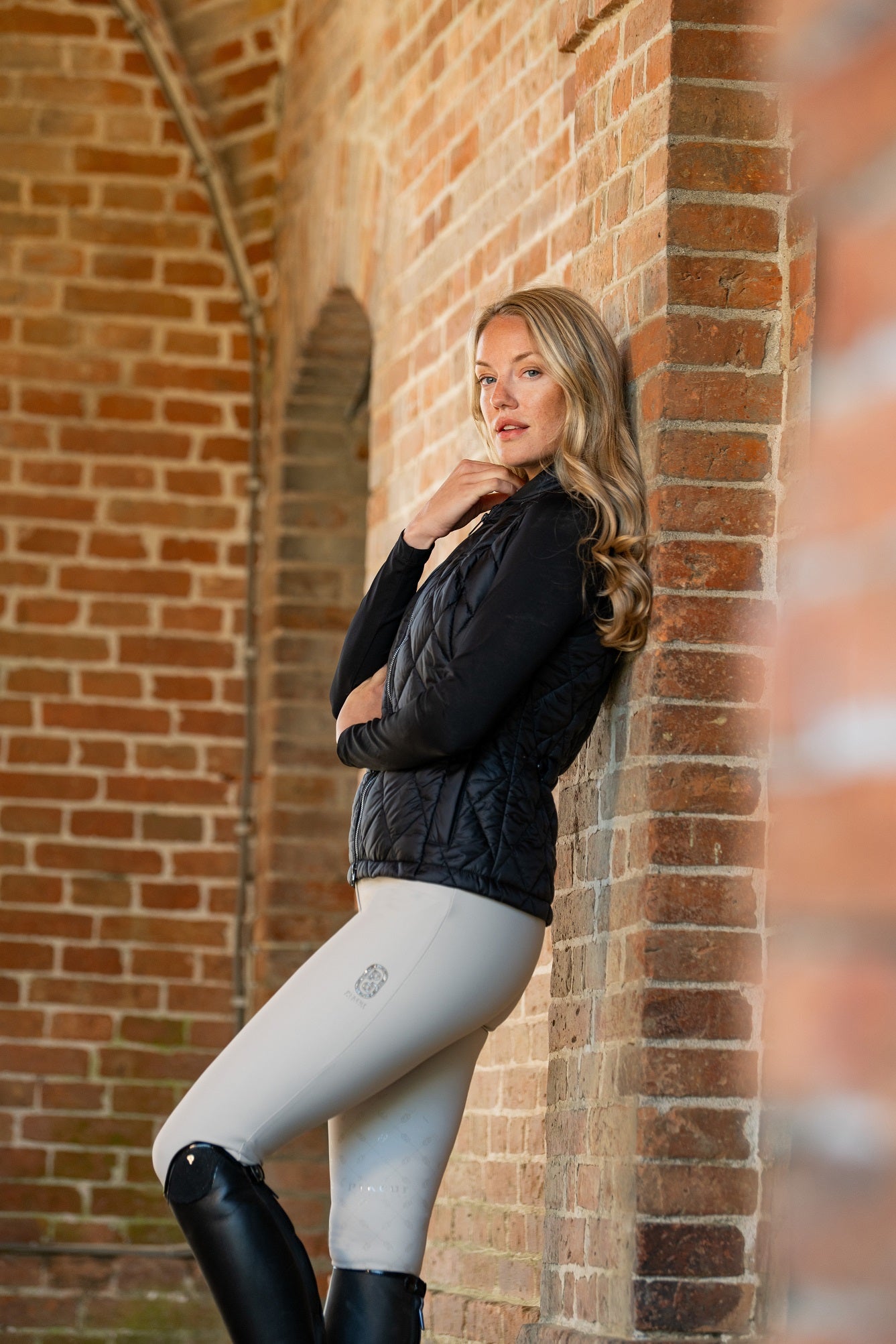 Woman in black jacket and gray leggings leaning against a brick wall.
