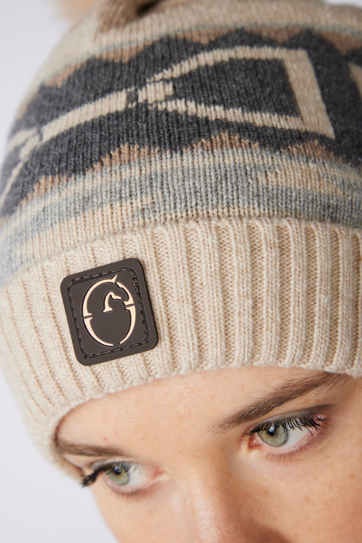 Close-up of a person wearing a knit beanie with a geometric pattern in beige, gray, and brown, featuring a black square patch on the brim with a white horse logo.