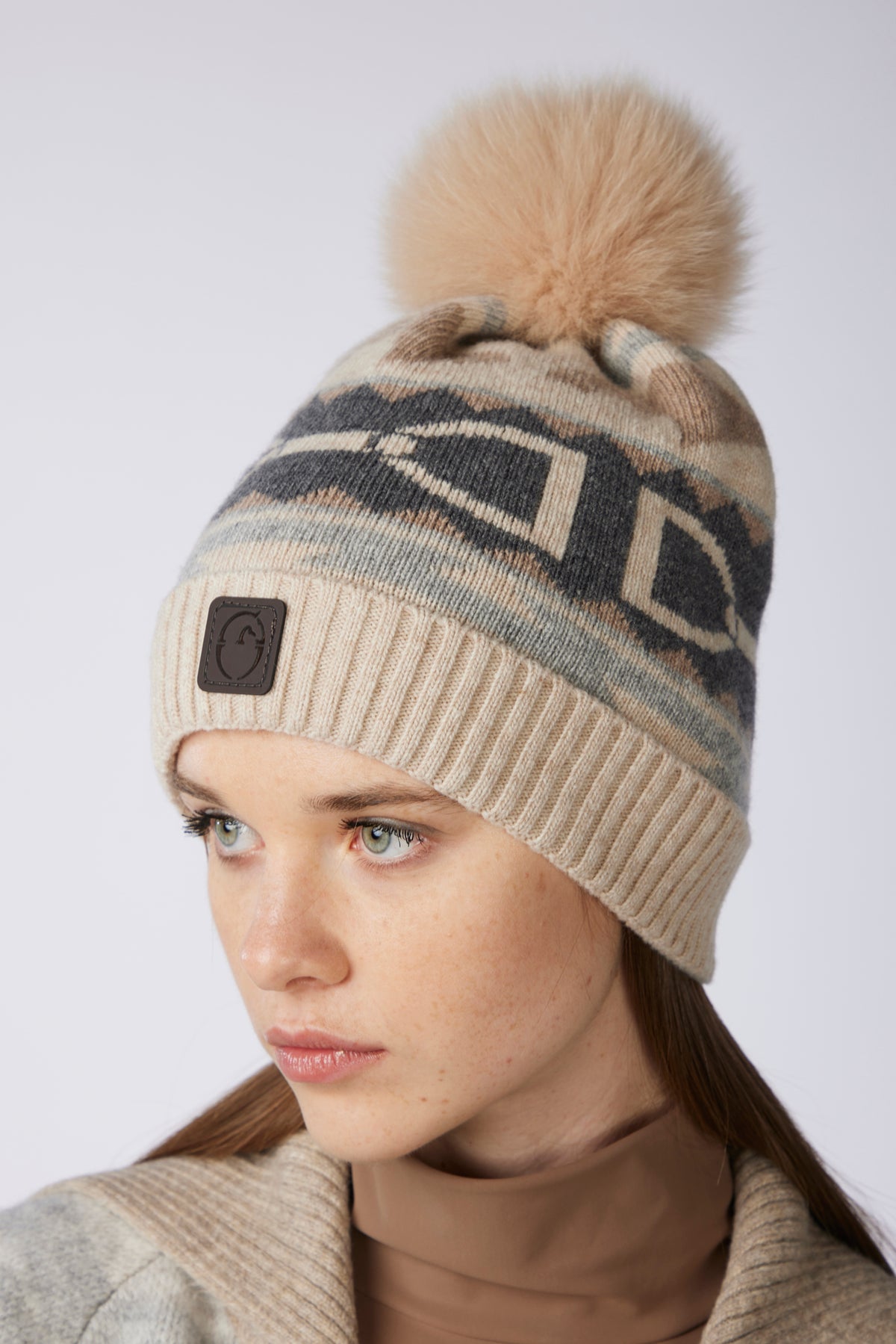 A close-up of a woman wearing a beige knit beanie with a large tan faux-fur pom-pom. The beanie features a geometric pattern in dark gray and beige, with a small dark brown square patch on the front.