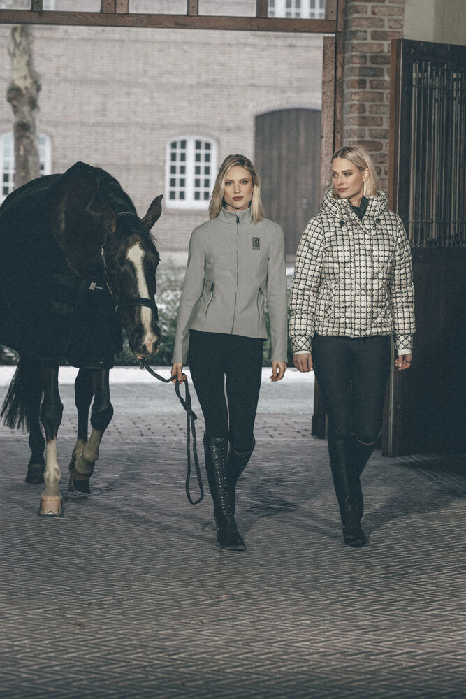 Two women walking with a horse in an outdoor stable setting, wearing equestrian apparel. One woman wears a light gray riding jacket with black pants and tall black boots, while the other wears a black-and-white checkered riding jacket with black pants and tall black boots.
