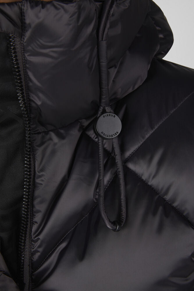 Close-up of a black puffer jacket with a drawstring hood and a round black brand tag on the cord. The zipper and quilted texture are visible against a light gray background.