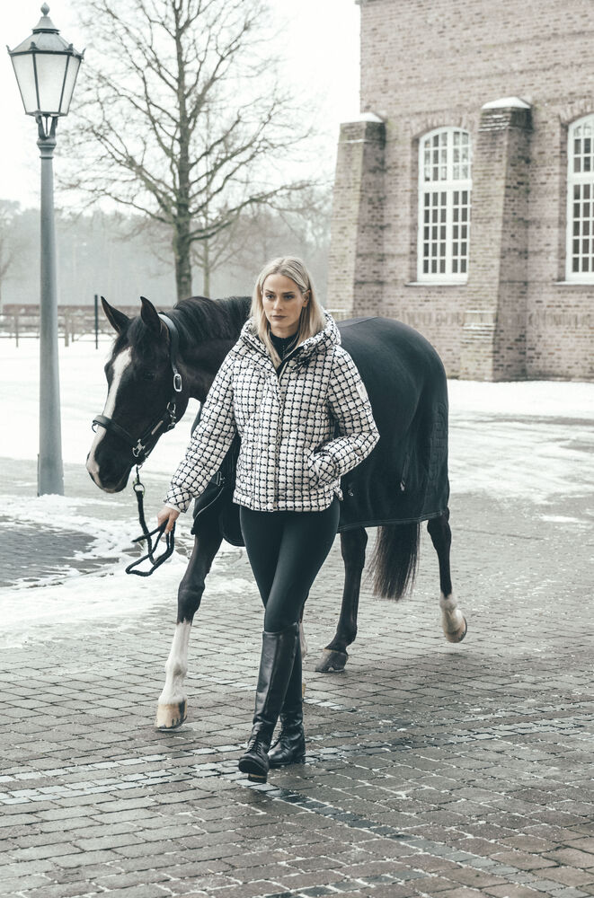 A woman walks with a black horse on a cobblestone street in front of a brick building. The woman is wearing a black and white checkered puffer jacket, black pants, and tall black boots, and holds the horse's reins.