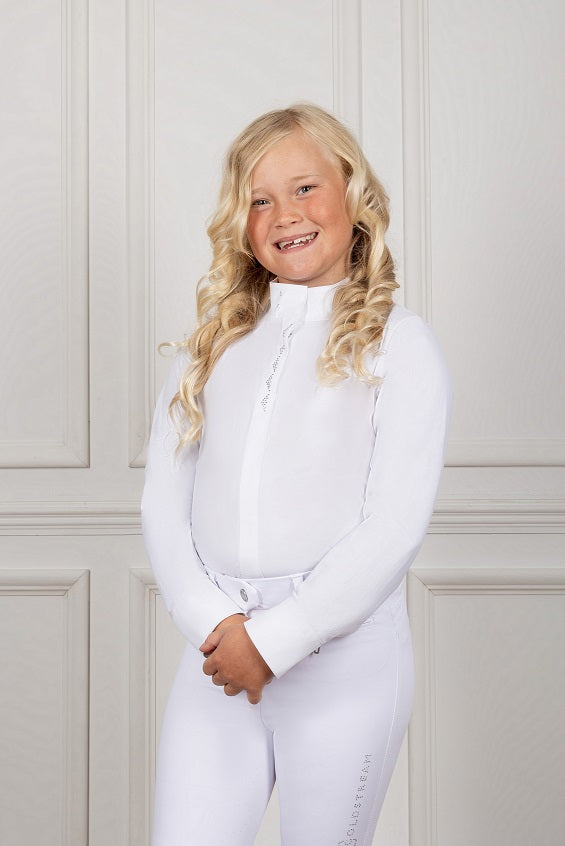 Young girl wearing a white equestrian outfit against a light-colored wall.
