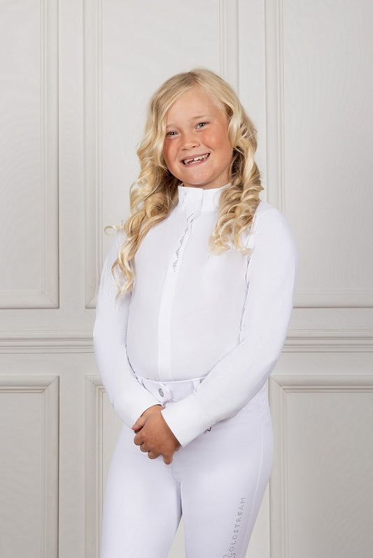 Young girl wearing a white equestrian outfit against a light-colored wall.