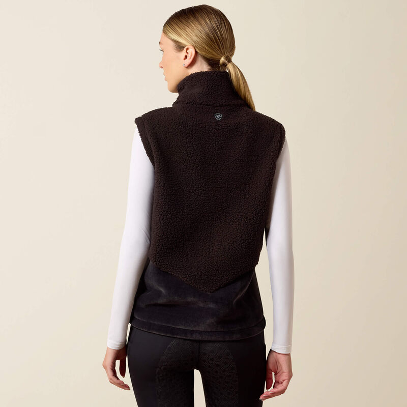 A woman shown from the back wearing a dark brown sherpa fleece vest over a white long-sleeve shirt and black leggings, against a plain beige studio background.