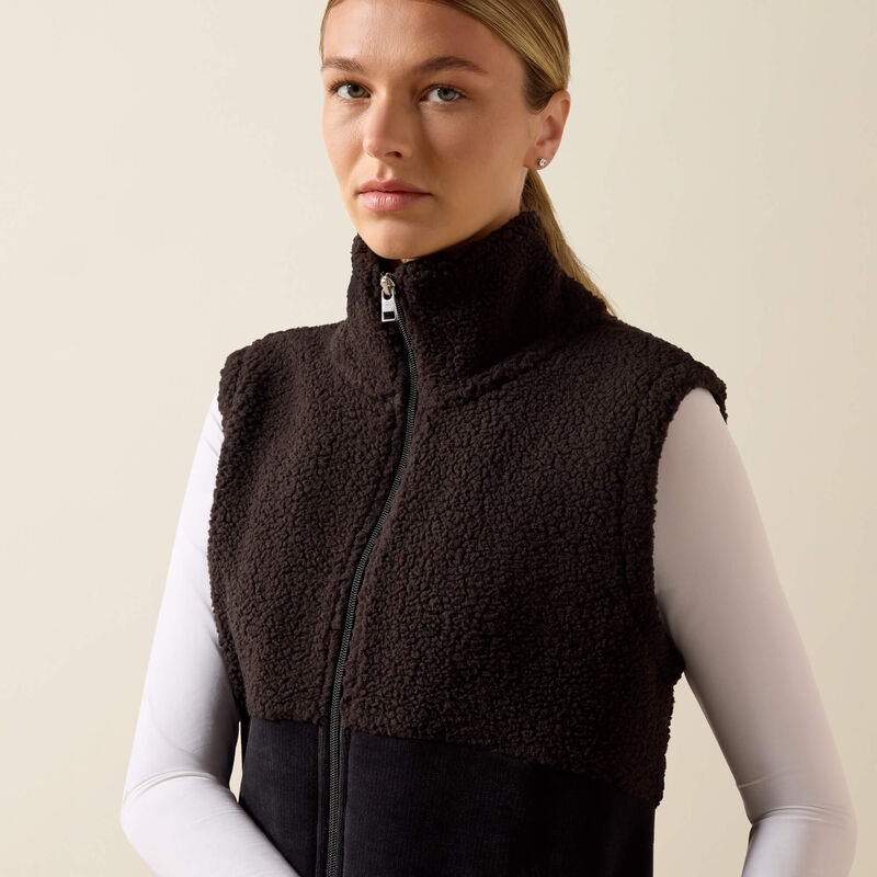 A woman wearing a dark brown sherpa fleece zip-up vest over a white long-sleeve top, photographed against a plain light beige studio background.