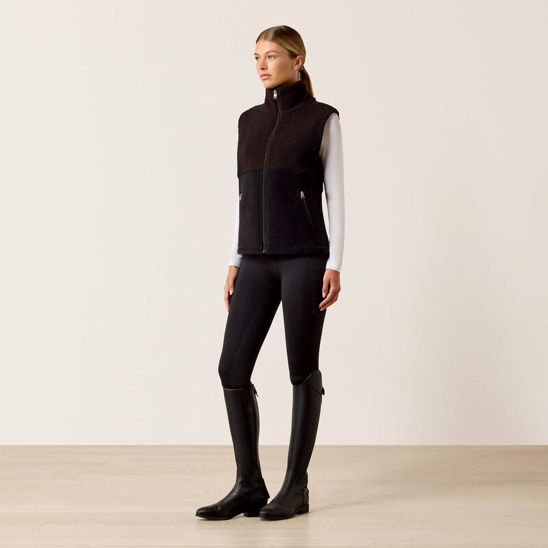 A woman stands in a studio wearing a sleeveless black and dark brown textured zip-front vest over a white long-sleeve top, black riding tights, and tall black riding boots against a plain light beige wall and pale wood floor.