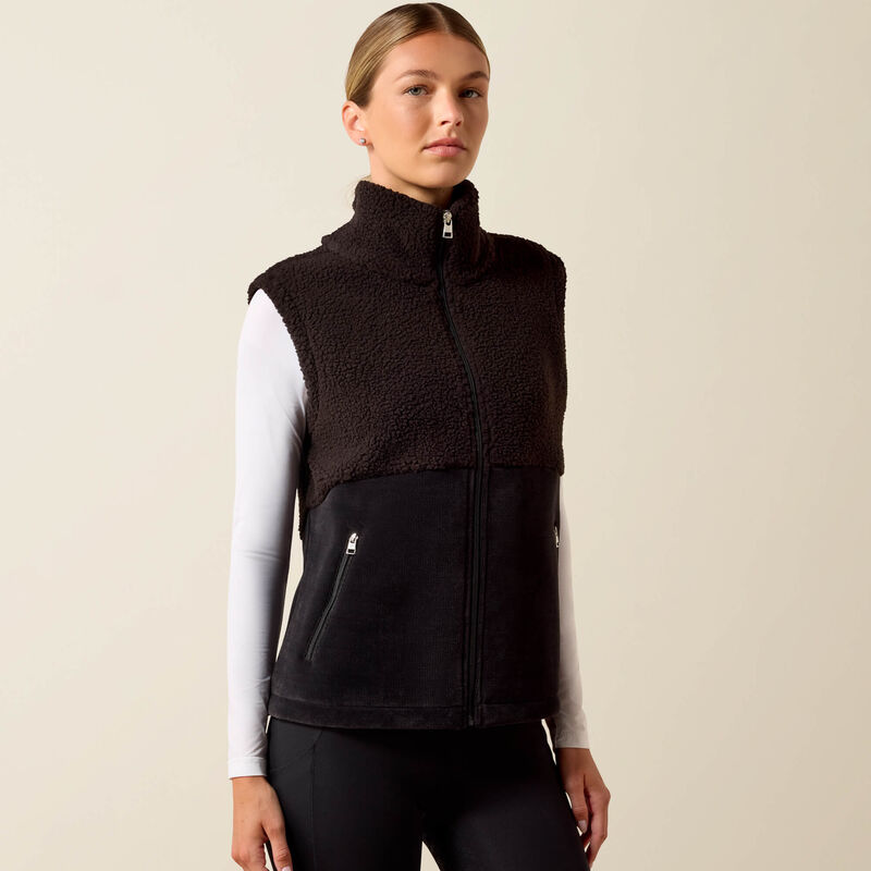 A woman modeling a sleeveless zip-front fleece vest with a dark brown sherpa upper section and a black lower section, worn over a white long-sleeve top and black leggings against a plain beige studio background.